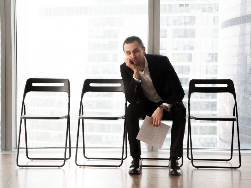 Bored young man in formal suit sitting on office chair in waiting room with document or resume in hand. Young job candidate or businessman waiting too long for interview or business meeting to start.