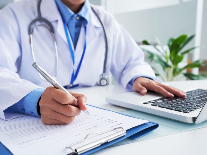 hands-unrecognizable-female-doctor-writing-form-typing-laptop-keyboard (1)